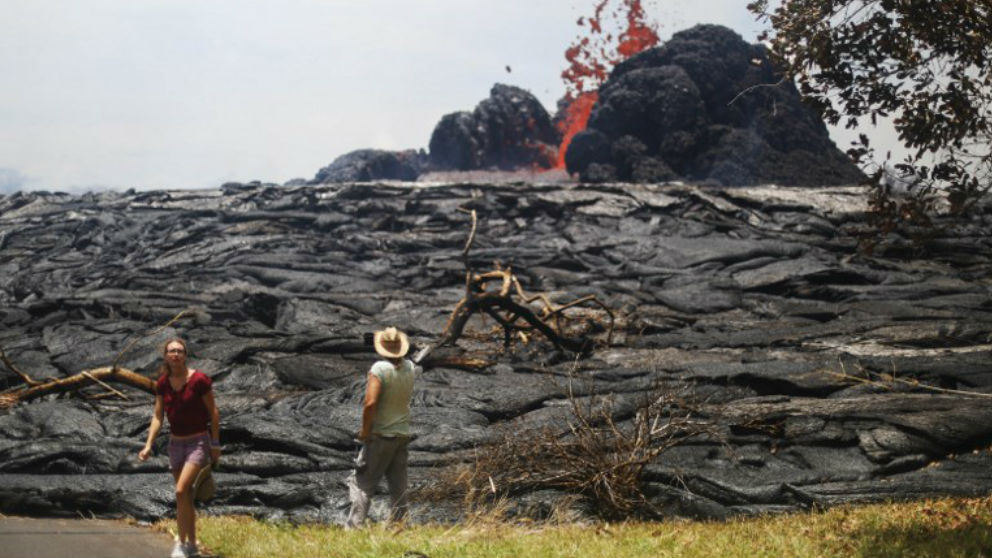Dos personas frente a un enorme muro de lava solidificada, detrás del cual corre un río de lava procedente del volcán Kilauea en Hawái. Foto: AFP