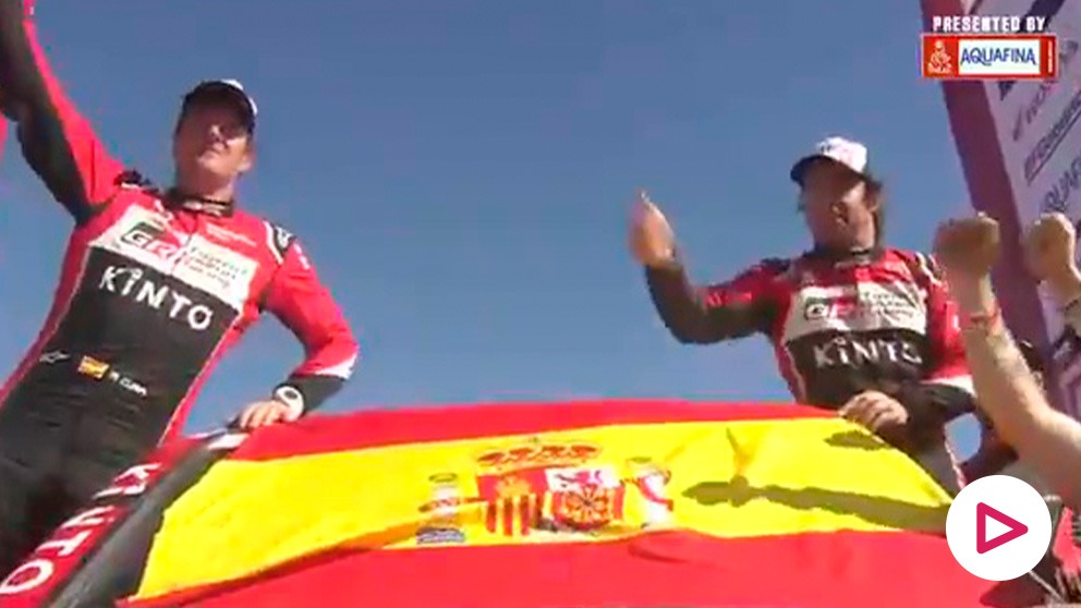 Fernando Alonso y Marc Coma saludan con una bandera de España posada en el murro del Toyota Hilux.