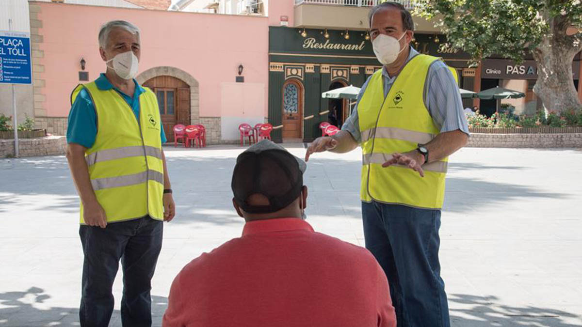 Dos voluntarios informan a un ciudadanos sobre el uso obligatorio de mascarillas en Alcarrás (Lérida).