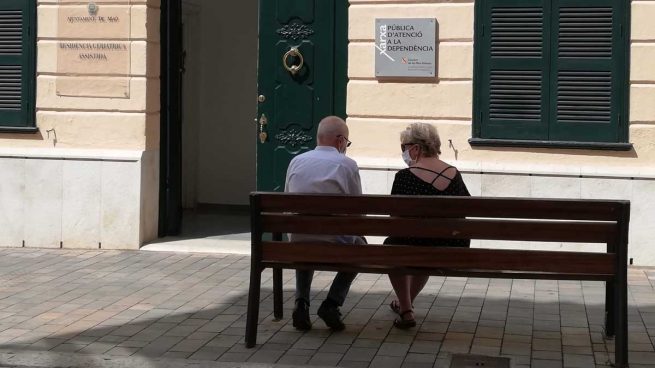Dos personas mayores conversan sentadas en un banco y con mascarilla.