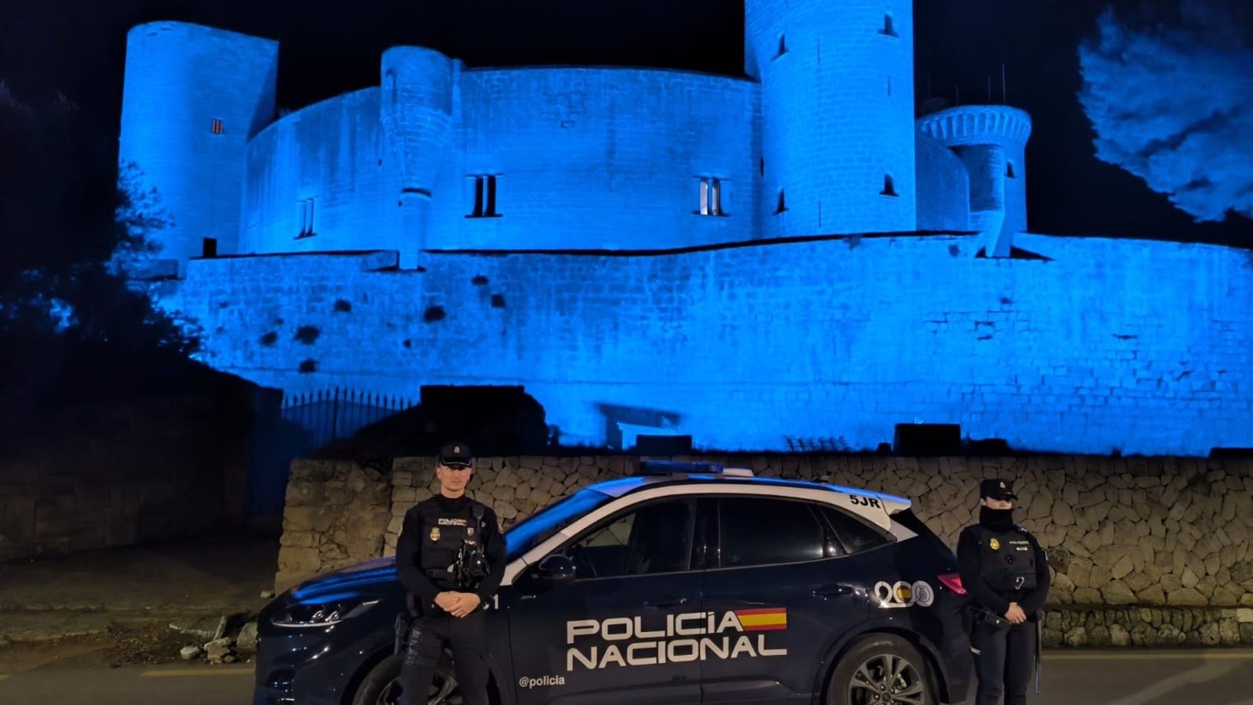 Agentes de la Policía Nacional, junto a un vehículo policial, y, tras ellos, el Castillo de Bellver, teñido de azul por el Bicentenario del Cuerpo.