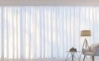 Salón con una silla, una mesita y gran ventanal con unas cortinas blancas.