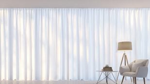 Salón con una silla, una mesita y gran ventanal con unas cortinas blancas.