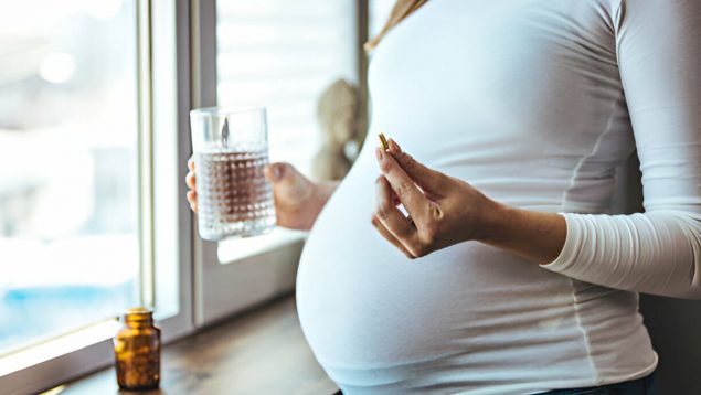 Mujer embarazada tiene una pastilla en la mano y un vaso de agua en la otra.