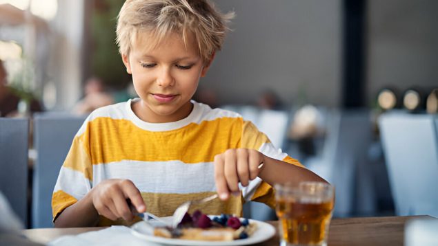 Niño rubio sonríe mientras corta su comida en un plato.