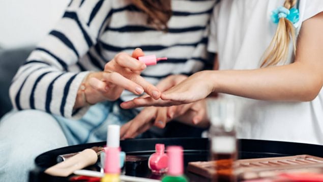 Madre pinta uñas a su hija con pintauñas abierto en la mesa.