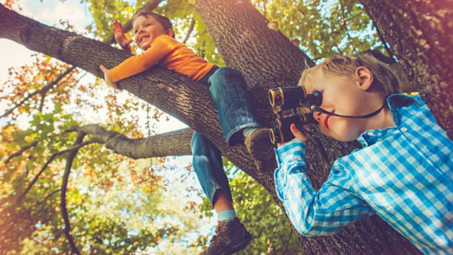 Dos niños jugando a explorar. Uno subido en un árbol y el otro con unos prismáticos.