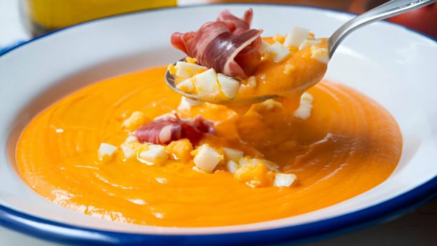 Foto de un plato de salmorejo cordobés con una cuchara que tiene jamón y huevo.