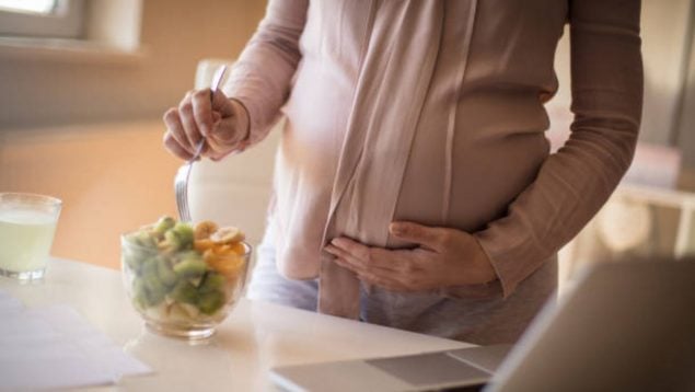 Mujer embarazada que se está preparando una ensalada en un bol de cristal mientras se toca la barriga.