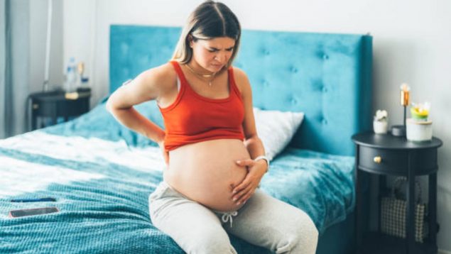 Mujer embarazada sentada en la cama mientra se toca la barriga.