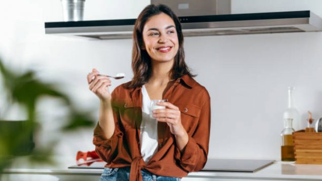 Mujer que come yogur, sujeta cuchara con una mano y sonríe.