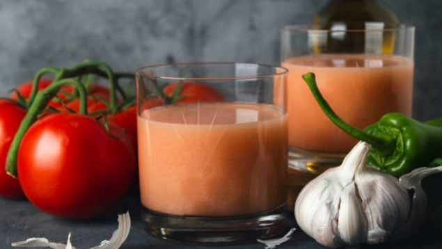 Foto de dos vasos de gazpacho llenos con tomates y ajos alrededor.