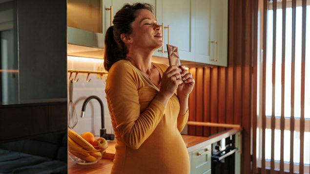 Foto de embarazada que sujeta una tableta de chocolate y sonríe antes de comerla.