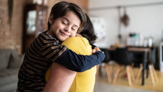 Foto de una madre con camiseta amarilla de la que se ve su espalda que lleva su hijo en brazos y que sonriente le abraza.