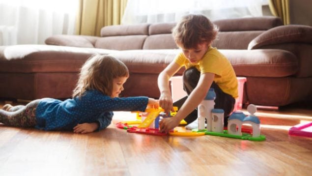 Un niño de rodillas y una niña tumbada en el suelo del salón de una casa mientras juegan con construcciones.