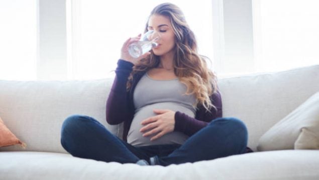 Foto de una mujer embarazada que estando sentada bebe un vaso de agua.