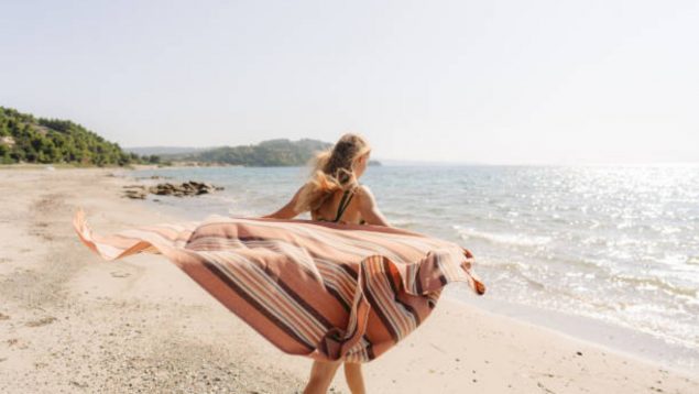 Foto de mujer de espaldas en la playa que camina sujetando la toalla que abierta está detrás de su espalda.