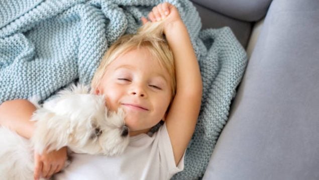 Foto de un niño rubio que está tumbado en un sofá sobre una colcha azul. Tiene los ojos medio cerrados, sonríe y a su lado se ve la cabeza de su perro.