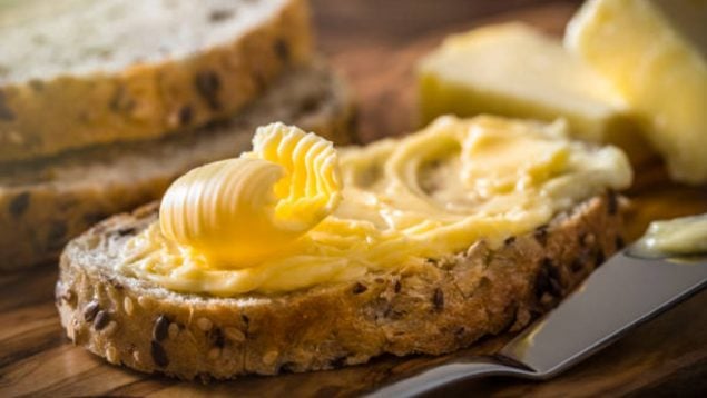 Foto de una rebanada de pan con semillas y encima tiene mantequilla extendida y al lado hay una espátula.