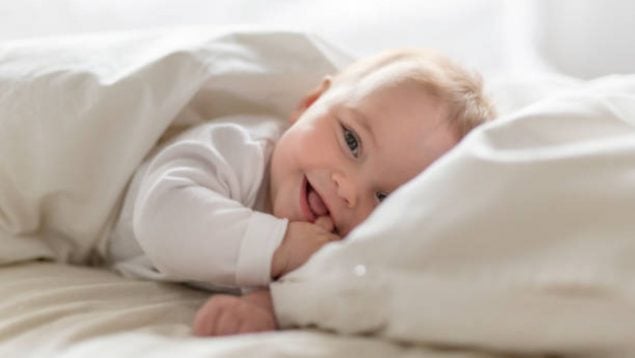 Foto de un bebé tapado y vestido de blanco que sonríe a cámara mientras está tumbado.