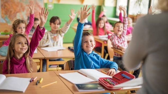 Foto de unos niños en una clase escolar que levantan la mano y sonríen.
