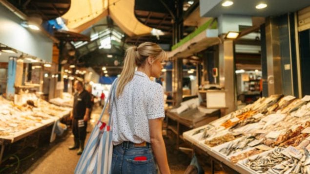 Foto de una mujer joven con coleta, blusa y tejanos, de espaldas a cámara que está mirando el pescado en una parada de un mercado.