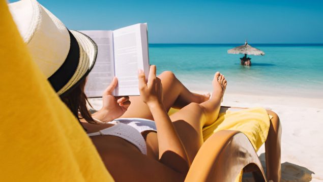 Foto de una mujer tumbada en una tumbona de playa con la orilla de fondo y está leyendo un libro.