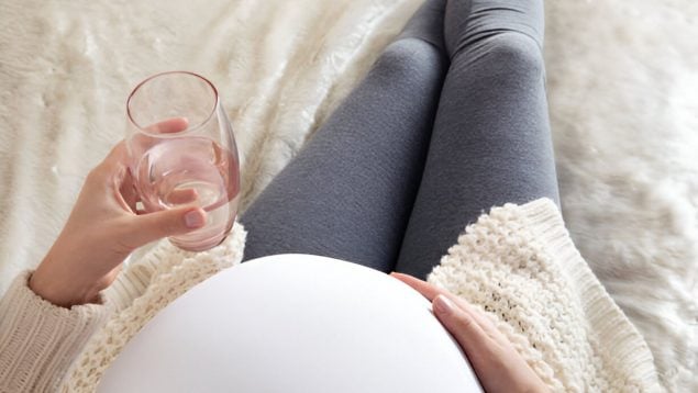 Foto de una mujer de la que se ve su barriga de embarazada mientras está sentada y sostiene un vaso de agua.