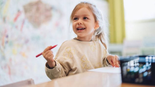 Foto de una niña pequeña que sentada en una clase tiene un lápiz en la mano y sonríe.