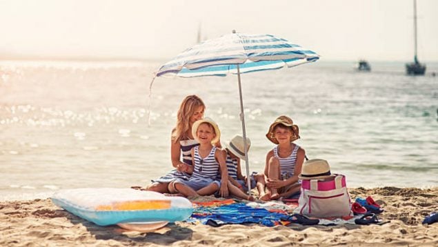 Foto de tres niños y una madre en la playa debajo de la sombrilla.