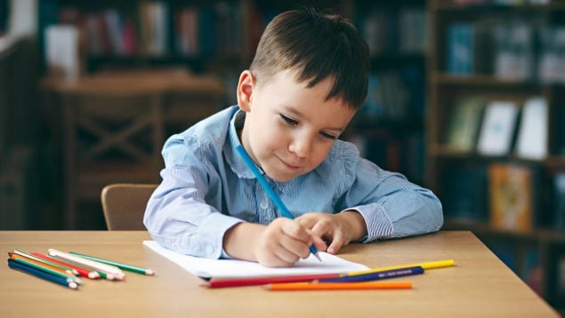 Niño pequeño escribe en una hoja con un lápiz de forma concentrada pero también está sonriendo.