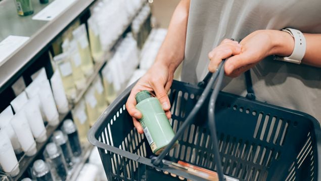 Foto de un cesto de supermercado y se ve la mano de una mujer que coloca una botella pequeña de champú de color verde.