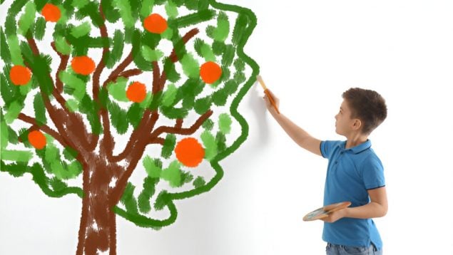 Niño con una paleta de pintor pintando un árbol grande con manzanas rojas sobre una pared blanca.