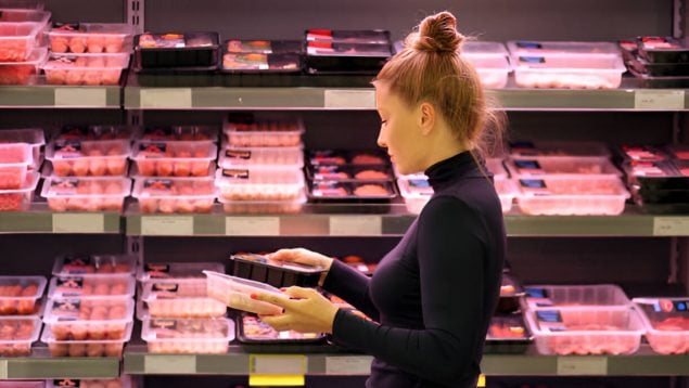 Foto de una mujer delante de una nevera de bandejas de carne. Llleva jersey negro y moño y sostiene dos bandejas de carne y las está mirando.