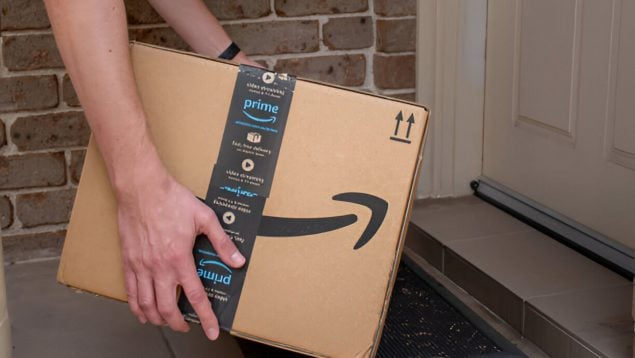 Foto de un repartidor de Amazon del que se ven los brazos que lleva una caja y está dejándola en el suelo delante de una puerta blanca.