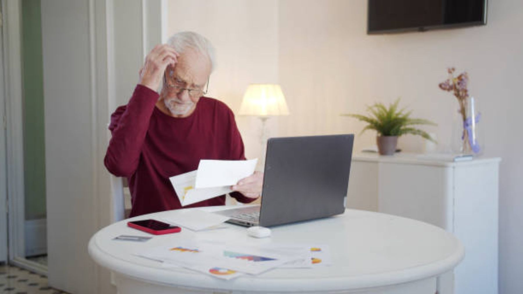 Hombre mayor leyendo documento frente a su ordenador.