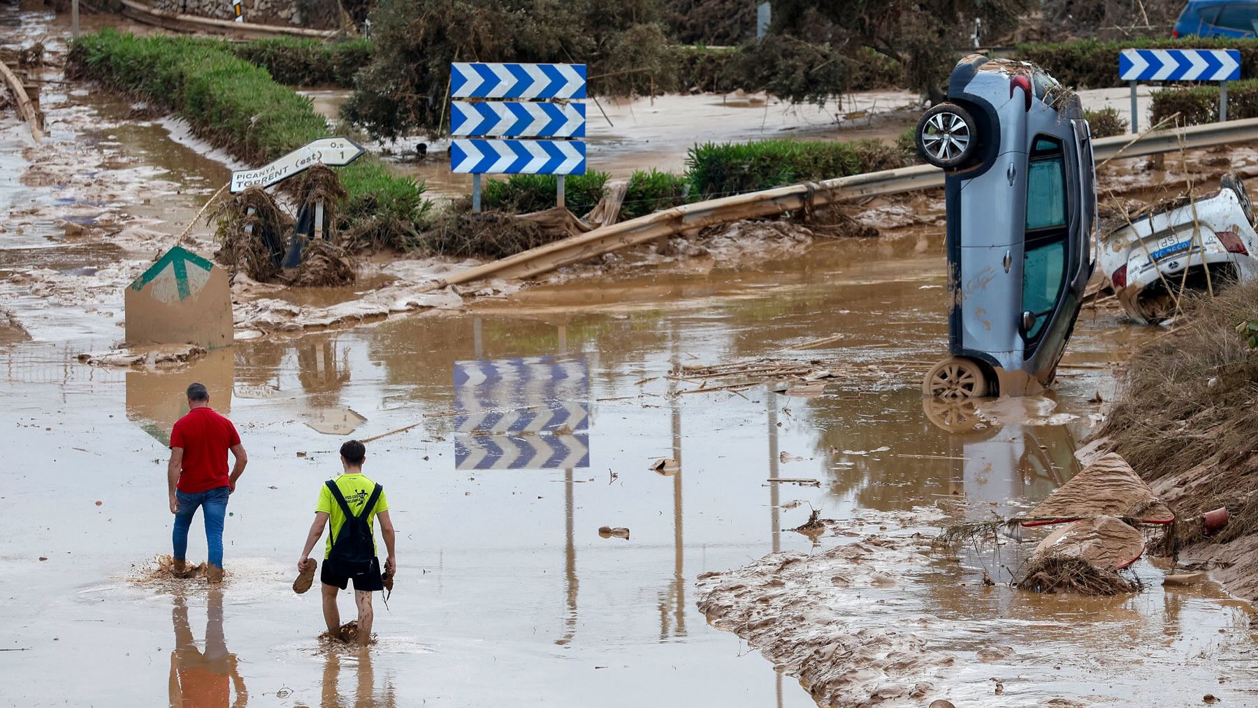 Dos personas se abren paso entre las aguas junto a un coche arrastrado por las riadas. (EFE)