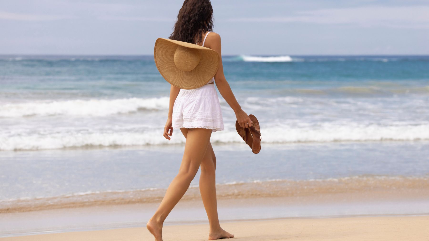 Una mujer anda por la playa con las sandalias en la mano.