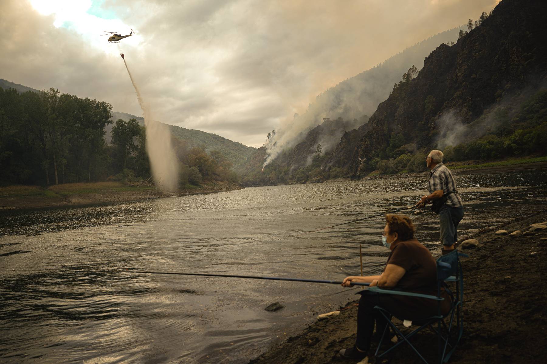 Un helicóptero recoge agua para ayudar con las labores de extinción del incendio, mientras una pareja pesca en Aguas Mestas, (Lugo). (Foto: Europa Press).