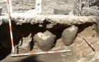 Dos vasijas de cerámica aparecen bajo una capa de tierra durante una excavación arqueológica, junto a los jalones que se usan para medir el yacimiento. Foto: Antequem