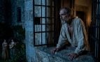 Don Quijote, mayor y con ropa de dormir, se asoma con atención desde una ventana con rejas de un castillo de piedra por la noche. En el jardín a lo lejos, en penumbra, dos doncellas con vestidos de época. Foto: representación con inteligencia artificial.