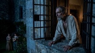 Don Quijote, mayor y con ropa de dormir, se asoma con atención desde una ventana con rejas de un castillo de piedra por la noche. En el jardín a lo lejos, en penumbra, dos doncellas con vestidos de época. Foto: representación con inteligencia artificial.