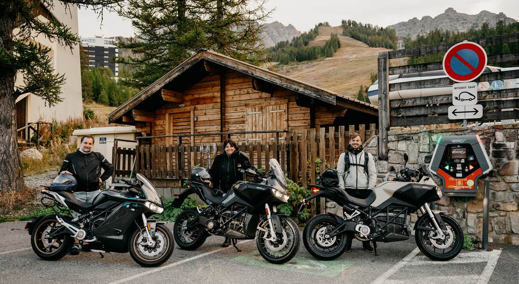 Tres pilotos junto a una SR/S, una DSR/X y una DS aparcadas al lado de un punto de recarga en los Alpes franceses