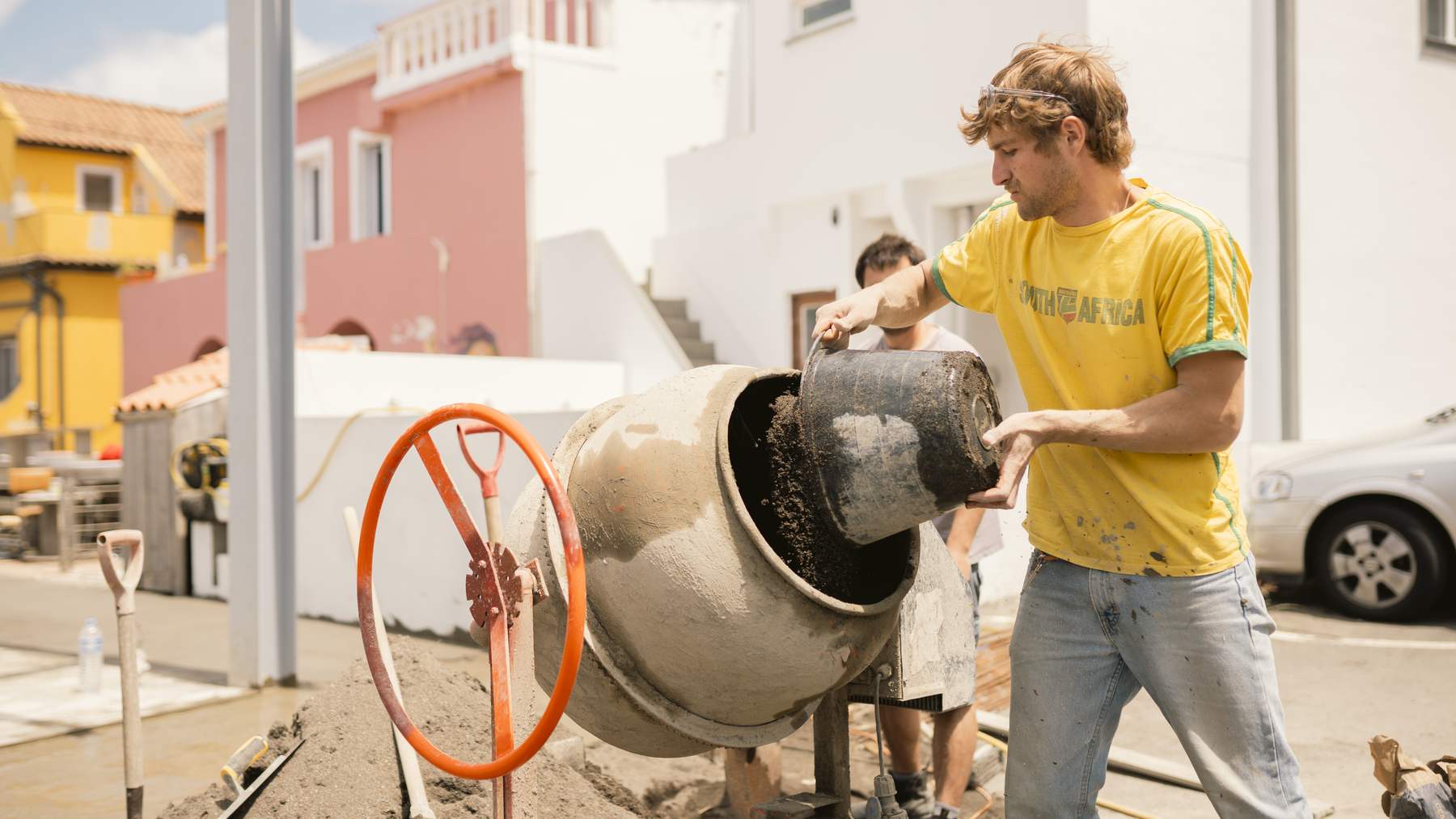 Un voluntario con camiseta amarilla echa un cubo de arena en una hormigonera junto a un edificio rosa en Fajã Grande