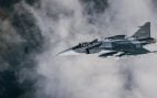 Caza Gripen E con insignias suecas, camuflaje gris y misiles bajo el ala, volando entre nubes