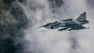 Caza Gripen E con insignias suecas, camuflaje gris y misiles bajo el ala, volando entre nubes
