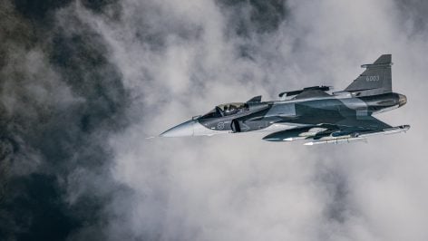 Caza Gripen E con insignias suecas, camuflaje gris y misiles bajo el ala, volando entre nubes
