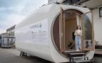 Casa diminuta de Vattenfall construida dentro de la góndola de un aerogenerador, con su gran abertura circular, las puertas de madera abiertas y una mujer en la entrada