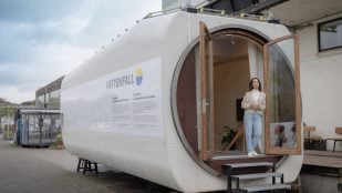 Casa diminuta de Vattenfall construida dentro de la góndola de un aerogenerador, con su gran abertura circular, las puertas de madera abiertas y una mujer en la entrada