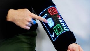 Un dedo toca la pantalla táctil de LED tejida en la manga de un jersey negro, que muestra un menú con iconos